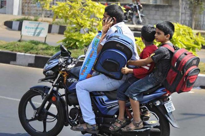 BA*****S of BHARAT - Shitwhole A man riding a motorcycle while talking on his phone, with two schoolchildren sitting behind him wearing backpacks on a city street.