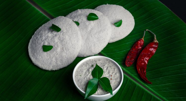 Three white idlis arranged on a banana leaf with a bowl of coconut chutney garnished with green leaves and two red dried chilies nearby