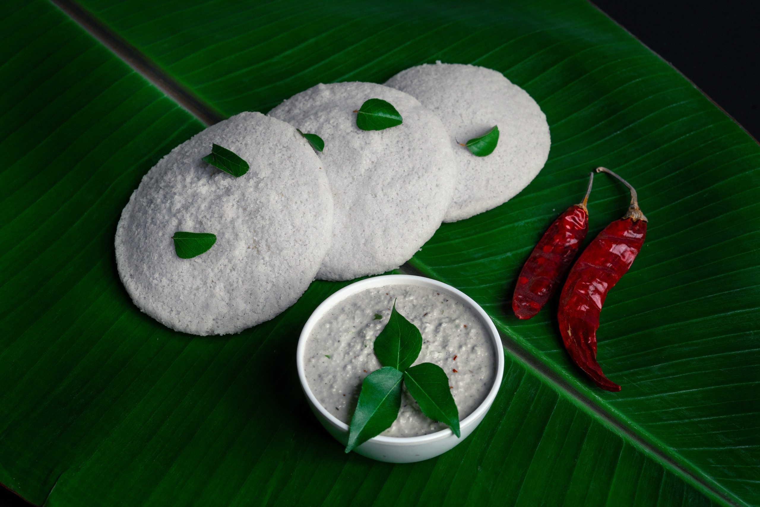 Three white idlis arranged on a banana leaf with a bowl of coconut chutney garnished with green leaves and two red dried chilies nearby