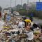 Workers sift through a massive pile of garbage on a city street, with a bus, cars, and a blue directional sign in the background.