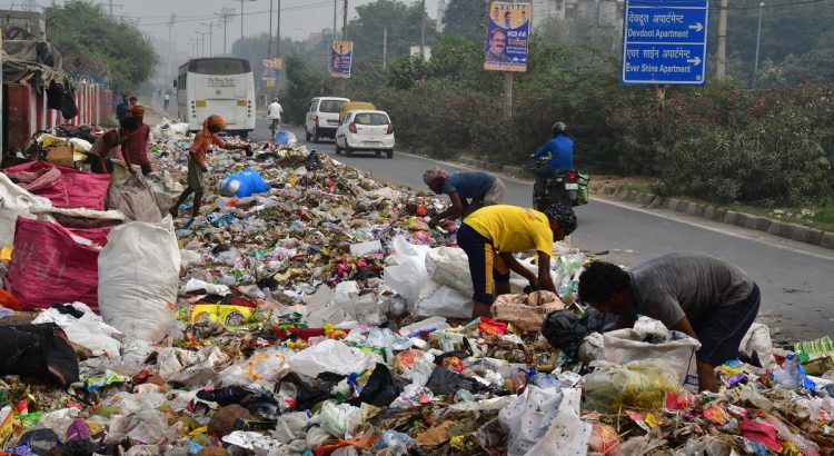 Workers sift through a massive pile of garbage on a city street, with a bus, cars, and a blue directional sign in the background.