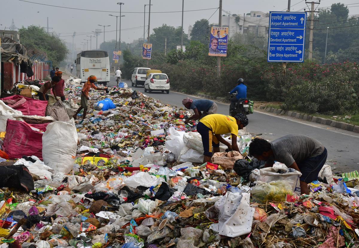 Workers sift through a massive pile of garbage on a city street, with a bus, cars, and a blue directional sign in the background.