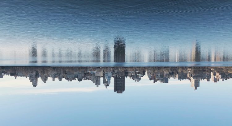 City skyline reflected in a calm river against a clear blue sky, with tall buildings along the waterfront.