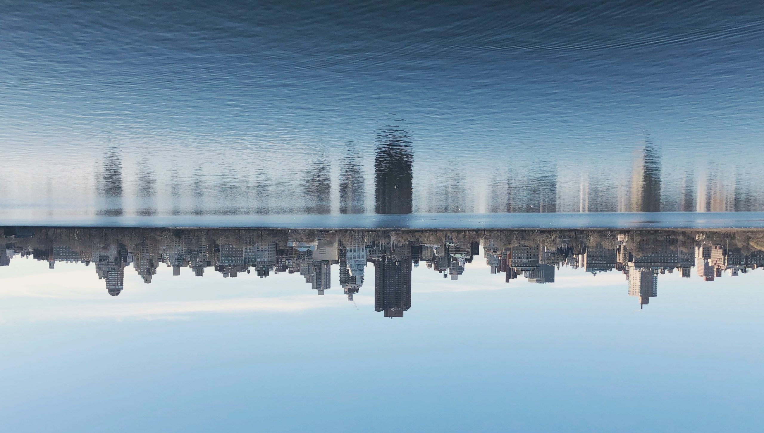 City skyline reflected in a calm river against a clear blue sky, with tall buildings along the waterfront.