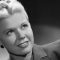Black-and-white portrait of a smiling woman with short blonde hair, looking upward to the left.