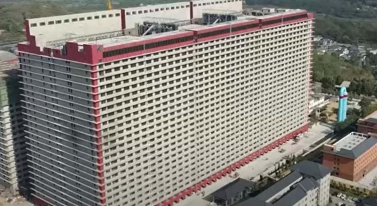 Aerial view of a large beige hotel with red trim, tall and rectangular, with a blue statue nearby and surrounding buildings.