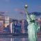 Statue of Liberty with the NYC skyline at dusk, lights reflecting on the water in front of tall buildings.