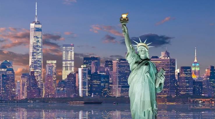 Statue of Liberty with the NYC skyline at dusk, lights reflecting on the water in front of tall buildings.