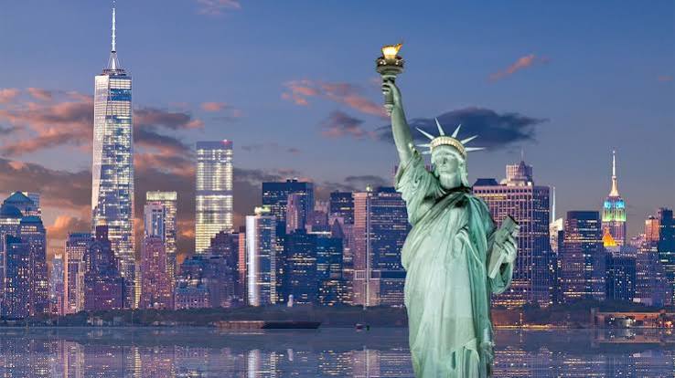 Statue of Liberty with the NYC skyline at dusk, lights reflecting on the water in front of tall buildings.