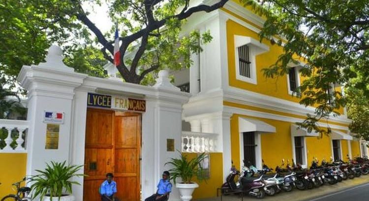 Yellow and white French lycée building with a large wooden gate, two guards sitting outside, and motorcycles along the sidewalk.