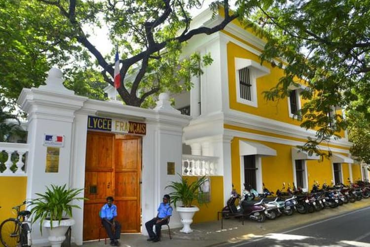 Yellow and white French lycée building with a large wooden gate, two guards sitting outside, and motorcycles along the sidewalk.